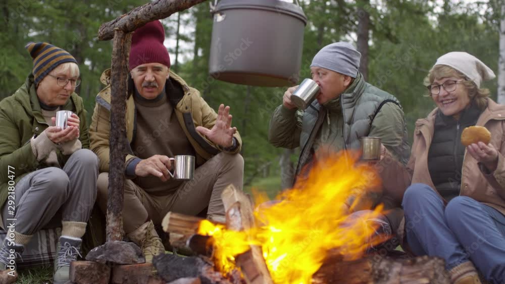 Cheerful senior men and women sitting beside campfire in forest, drinking hot tea, eating buns and speaking about something Cheerful senior men and women sitting beside campfire in forest, drinking hot tea, eating buns and speaking about something