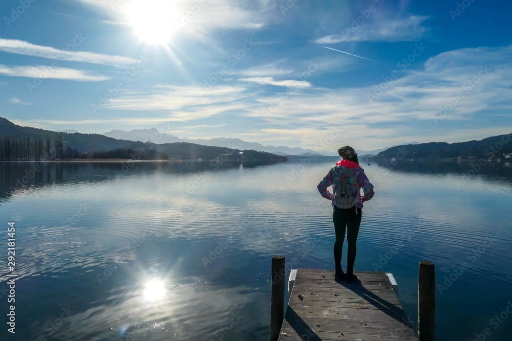 Girl wearing a jacket and beanie stands at the end of a promenade on the lake. Soft reflections of the clouds in the lake. Clear but cloudy day. High mountains in the back Calmness and relaxation