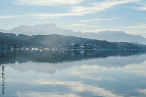 A view on a lake and Alps in the back. The calm surface of the lake is reflecting the mountains, sunbeams and clouds. Little city located at the lake side. Church with bell tower in the middle