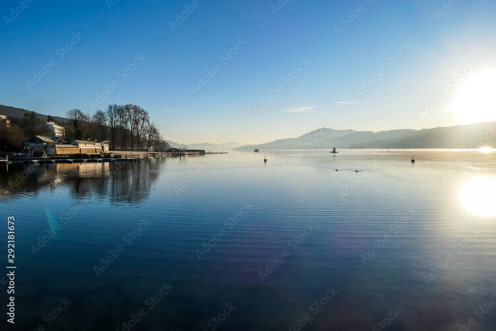 A view on a lake and Alps in the back. The calm surface of the lake is reflecting the mountains, sunbeams and clouds. Clear and sunny day. Calm and relaxed feeling. Few ducks crossing the lake