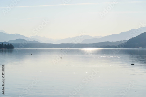 A view on a lake and Alps in the back. The calm surface of the lake is reflecting the mountains, sunbeams and clouds. Clear and sunny day. Calm and relaxed feeling. Few ducks crossing the lake