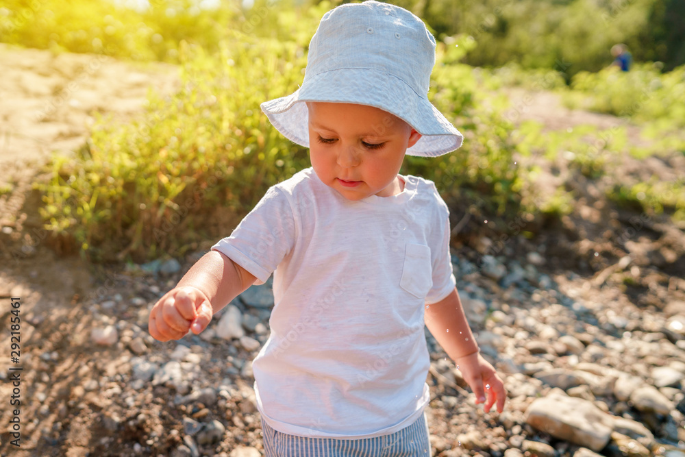 Portrait of playful small little boy child playing with rocks and water by the lake or river in sunny day wearing hat in nature Portrait of playful small little boy child playing with rocks and water by the lake or river in sunny day wearing hat in nature