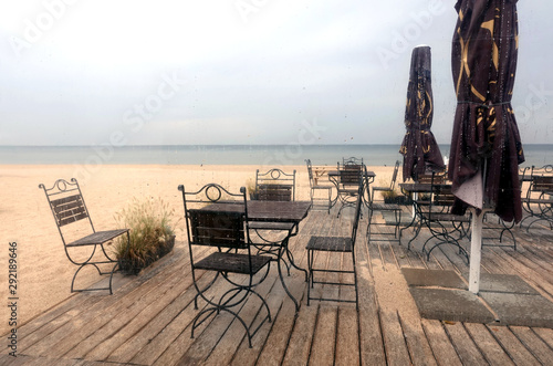 Several wooden tables and chairs on timber platform on empty seaside cafe on Jurmala beach in low season on rainy day view across glass