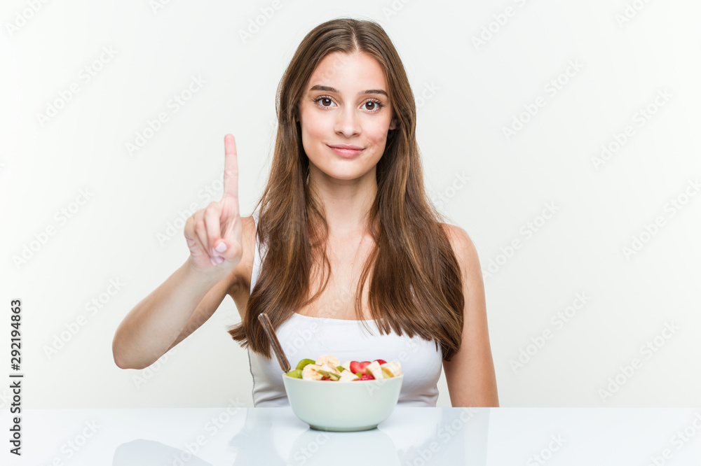 Young caucasian woman eating fruit bowl showing number one with finger. Young caucasian woman eating fruit bowl showing number one with finger.