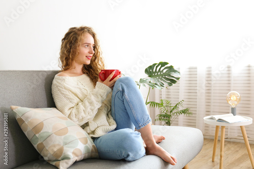 Young beautiful woman wearing white sweater on grey textile sofa at home. Attractive slim female in domestic situation, resting on couch in her lofty apartment. Background, copy space, close up.