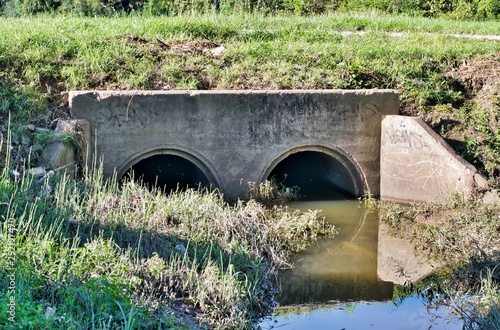 Culvert with twin concrete drains under an unpaved road in Humble, Texas. Dirty polluted water is stagnant in the drainage ditch with some trash lying in the grass. City pollution is getting worse. photo