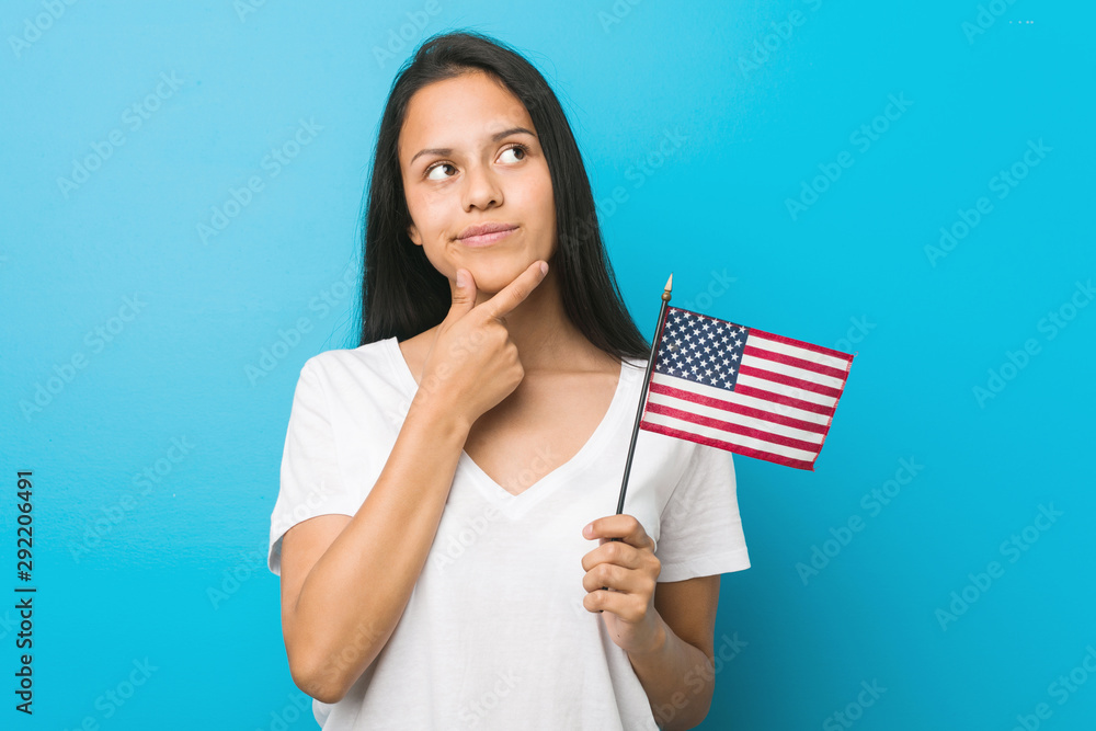 Young hispanic woman holding a united states flag looking sideways with doubtful and skeptical expression. Young hispanic woman holding a united states flag looking sideways with doubtful and skeptical expression.