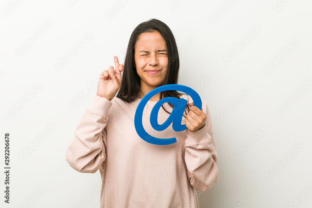 Young hispanic woman holding an at icon crossing fingers for having luck Young hispanic woman holding an at icon crossing fingers for having luck