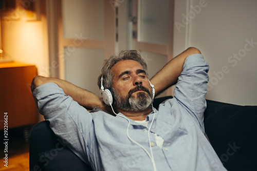 mid aged man relaxed in sofa listening music on headphones in his home