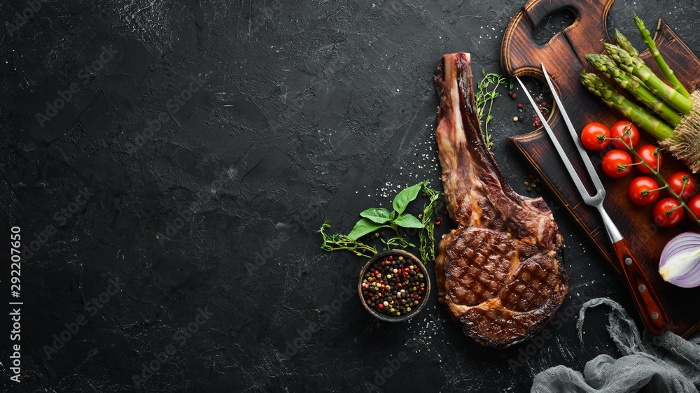 Grilled cowboy beef steak, herbs and spices on a black stone background. Barbecue. Top view. Free space for text. Grilled cowboy beef steak, herbs and spices on a black stone background. Barbecue. Top view. Free space for text.
