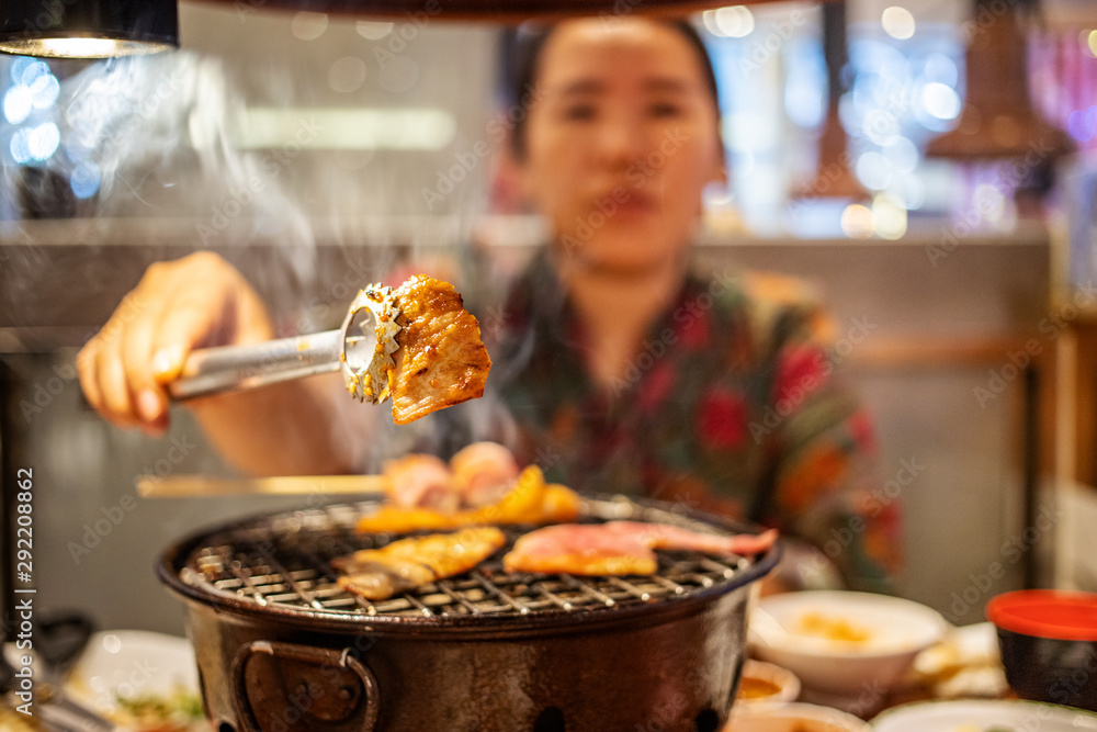 Woman using tong gripping a piece of sliced grilled pork over stove in Japanese restaurant
