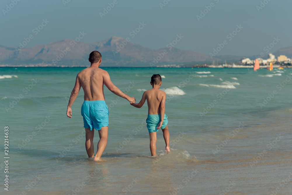 Dad and son in similar swimming shorts walking along beautiful seashore. They holding hands and enjoying their perfect vacations. Dad and son in similar swimming shorts walking along beautiful seashore. They holding hands and enjoying their perfect vacations.