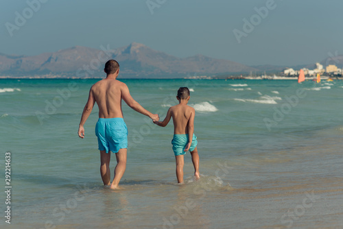 Dad and son in similar swimming shorts walking along beautiful seashore. They holding hands and enjoying their perfect vacations.