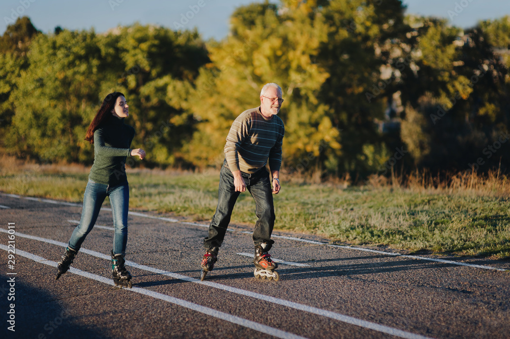 Fototapeta premium Parents and children spend time together. Older generation and sport. Active lifestyle of the pensioner. Happy old man riding on rollers with his daughter on the road in autumn park.