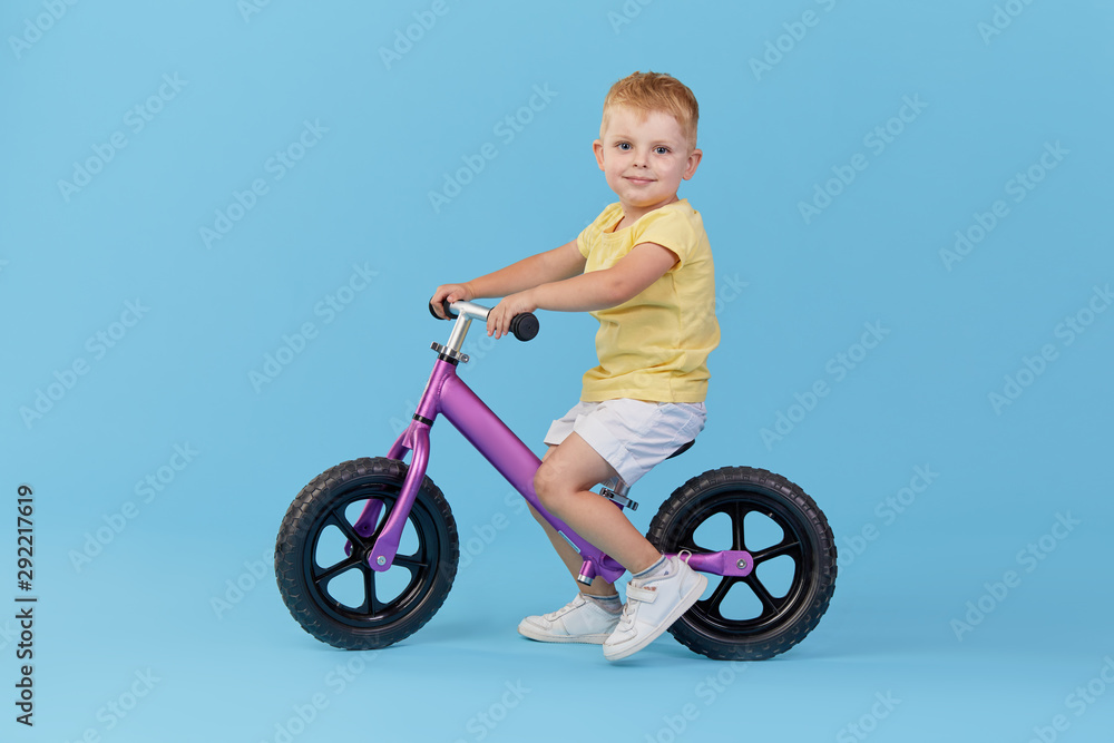 Little child boy learns to keep balance while riding a bicycle. Stylish child in yellow T-shirt on blue background Little child boy learns to keep balance while riding a bicycle. Stylish child in yellow T-shirt on blue background