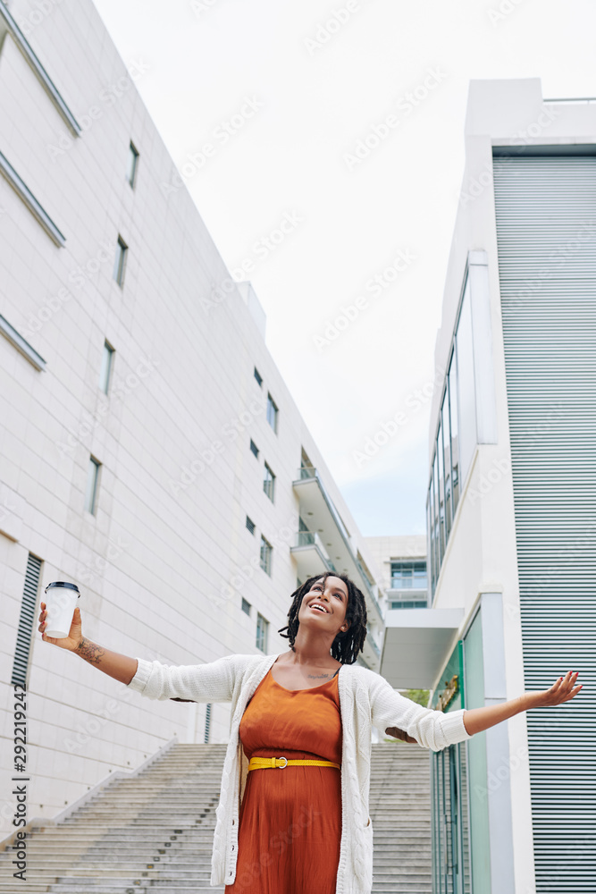 African happy woman in orange dress drinking coffee while standing outdoors and enjoying the good weather African happy woman in orange dress drinking coffee while standing outdoors and enjoying the good weather
