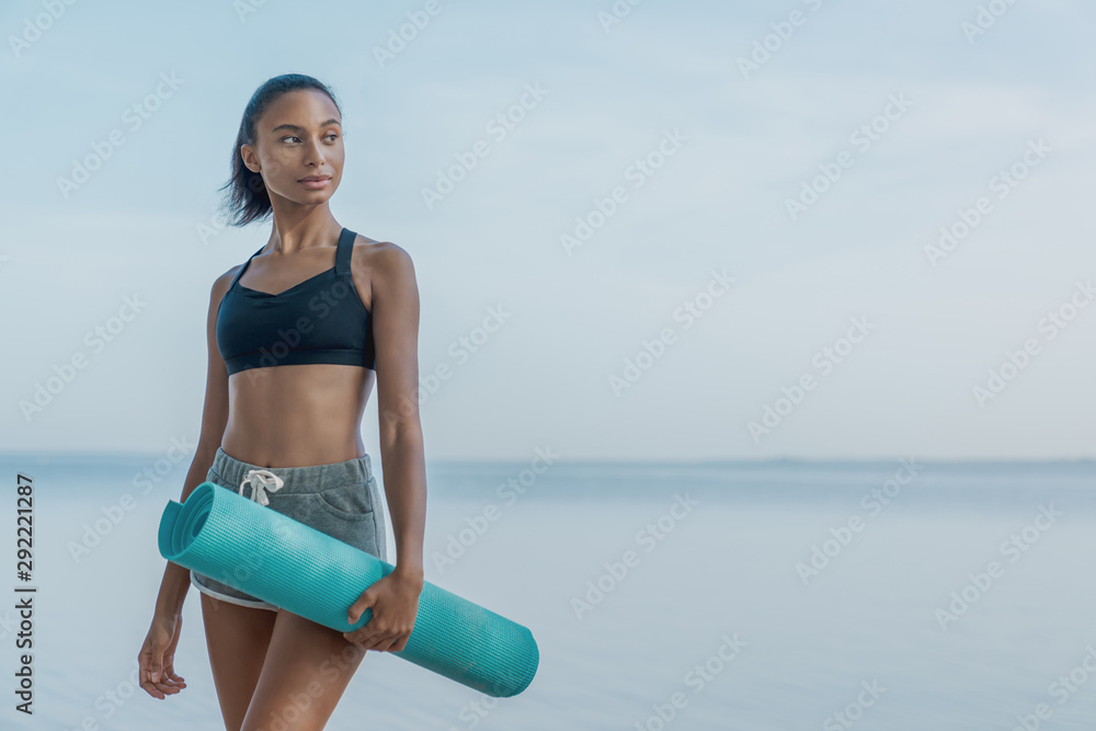 Smiling woman athlete in fitness clothes relaxing after workout with mat in  hand standing on the beach Stock Photo | Adobe Stock