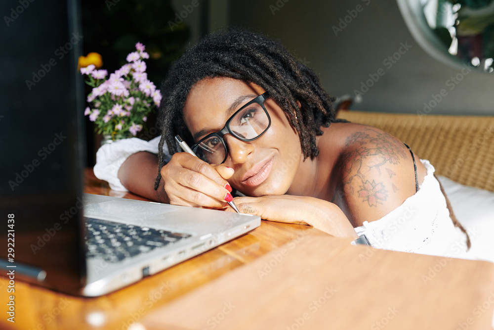 Portrait of African woman in eyeglasses looking at camera while lying at the table in front of laptop and writing her plans Portrait of African woman in eyeglasses looking at camera while lying at the table in front of laptop and writing her plans