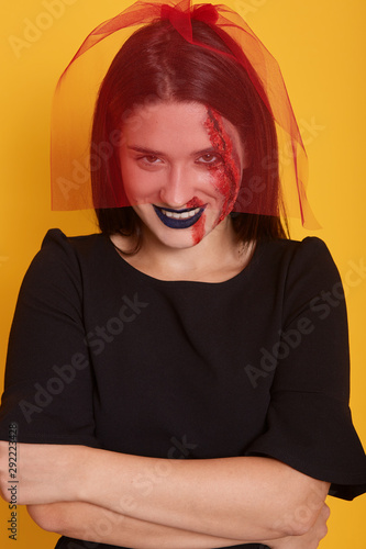 Front view of devil woman standing in nervous pose with folded hands isolated over yellow studio background, wearing red veil and black dress, has dangerous facial expression. Halloween concept.