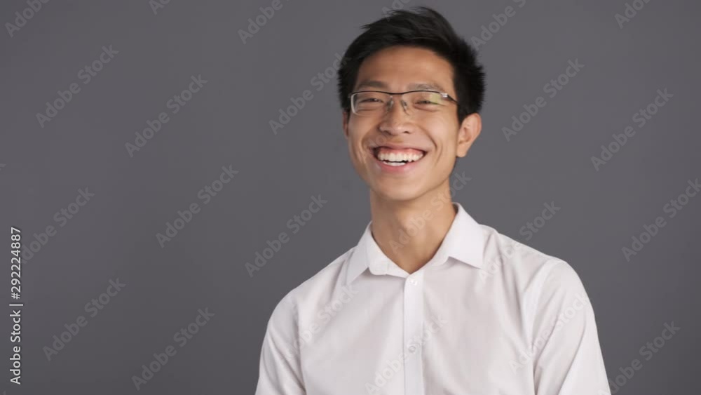Young cheerful asian man in eyeglasses joyfully looking in camera over gray background Young cheerful asian man in eyeglasses joyfully looking in camera over gray background