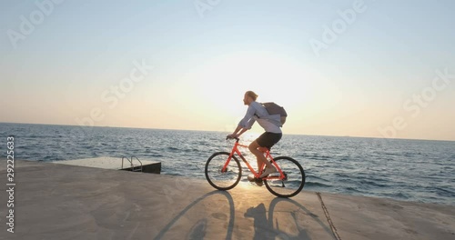Young handsome male in casual wear ride on the colorful bicycle on the morning beach against beautiful sunset and the sea 