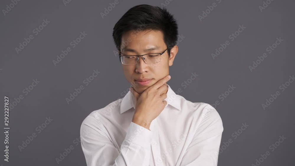 Young thoughtful asian man in eyeglasses holding thinking and waving head no gesture on camera over gray background Young thoughtful asian man in eyeglasses holding thinking and waving head no gesture on camera over gray background