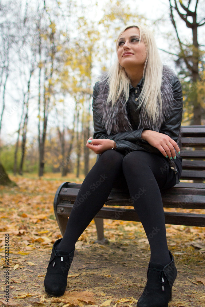 2017.10.21, Moscow, Russia. A young attractive woman wearing fur black jacket holding book sitting on the bench in falling park. Walking around autumn park. 2017.10.21, Moscow, Russia. A young attractive woman wearing fur black jacket holding book sitting on the bench in falling park. Walking around autumn park.