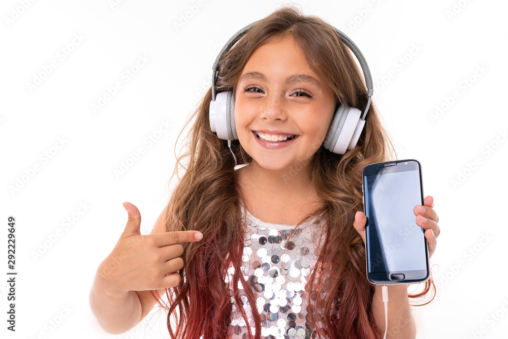 Beautiful girl in glittery dress, with big white headphones listening to music and showing screen of black smartphone and pointing at it isolated Beautiful girl in glittery dress, with big white headphones listening to music and showing screen of black smartphone and pointing at it isolated