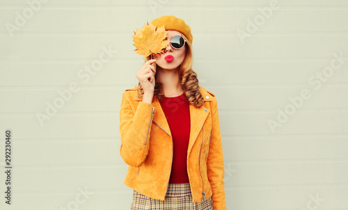 Autumn portrait woman holding in her hands yellow maple leaves hiding her eye, girl blowing red lips sending sweet air kiss over gray wall background