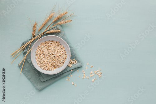Bowl of dry oat flakes with ears of wheat on light blue background. Cooking oats porridge concept