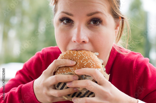 Close up portrait of hungry girl is eating fast street junk food. Young woman biting tasty yummy burger, hamburger outdoor. Extra calories, overeating. Bad dangerous habit, unhealthy lifestyle.