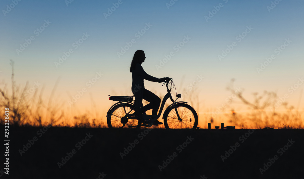 Silhouette of a young girl on the e-bike or electric bicycle on the sunset background. Country style. Transportation in the village. Copy space. Female. Travel.