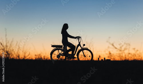 Silhouette of a young girl on the e-bike or electric bicycle on the sunset background. Country style. Transportation in the village. Copy space. Female. Travel.