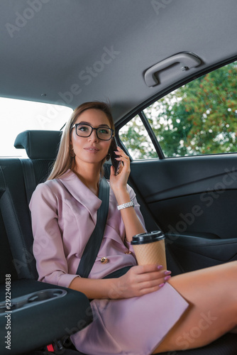 Beautiful business lady woman rides passenger compartment passenger car VIP taxi, fastens smartphone her hands, rings, listens road, important call from secretary. Long hair, cup coffee tea drink. © byswat