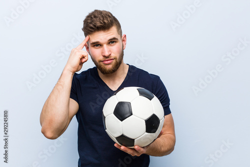 Young caucasian man holding a soccer ball pointing his temple with finger, thinking, focused on a task.