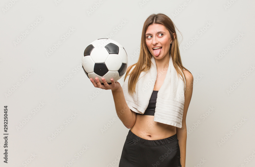 Young fitness russian woman funnny and friendly showing tongue. Holding a soccer ball.