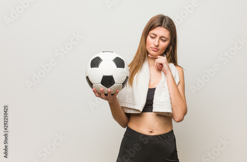 Young fitness russian woman doubting and confused. Holding a soccer ball.