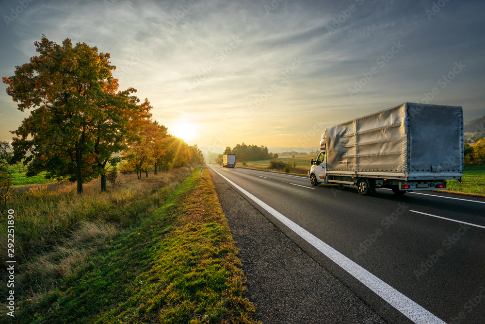 Delivery van and truck driving on the asphalt road towards sunset around line of trees in misty autumnal landscape