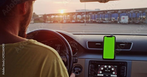 Close-up young man inside car using mounted smartphone navigator on greenscreen mock-up display driving at sunset in the city.