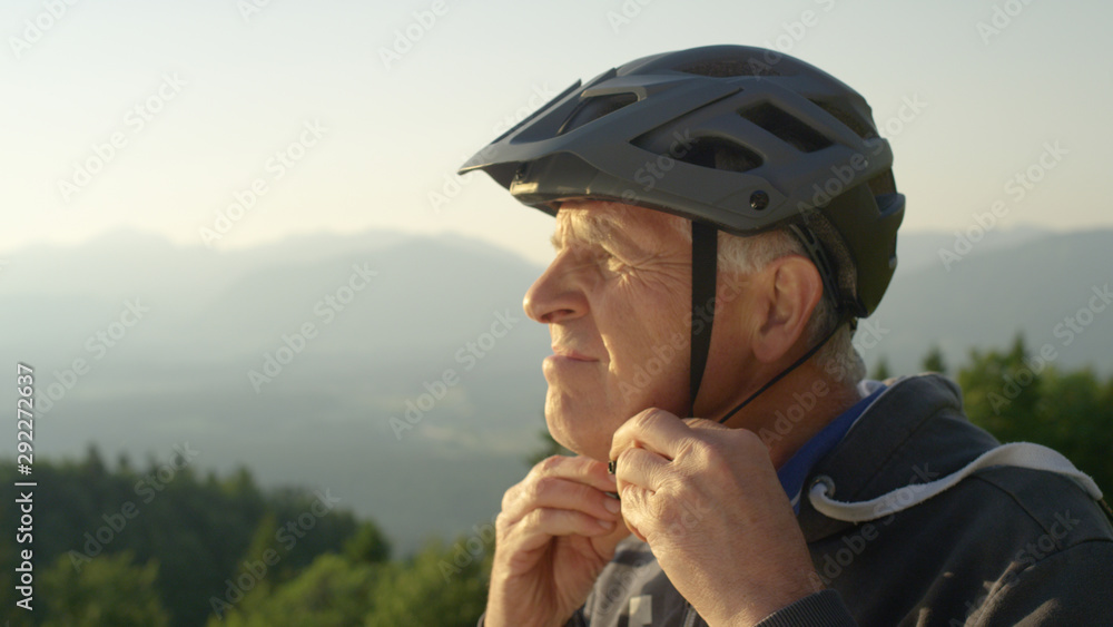 CLOSE UP: Focused senior man straps up his helmet before an evening bicycle ride - obrazy, fototapety, plakaty 