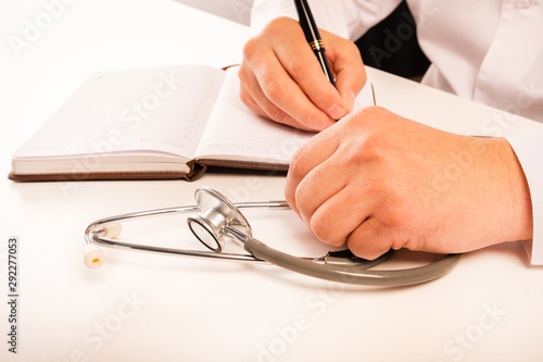 The doctor sits in an office at a white table, makes notes in a notebook, a stethoscope lies nearby, view from above. Close up