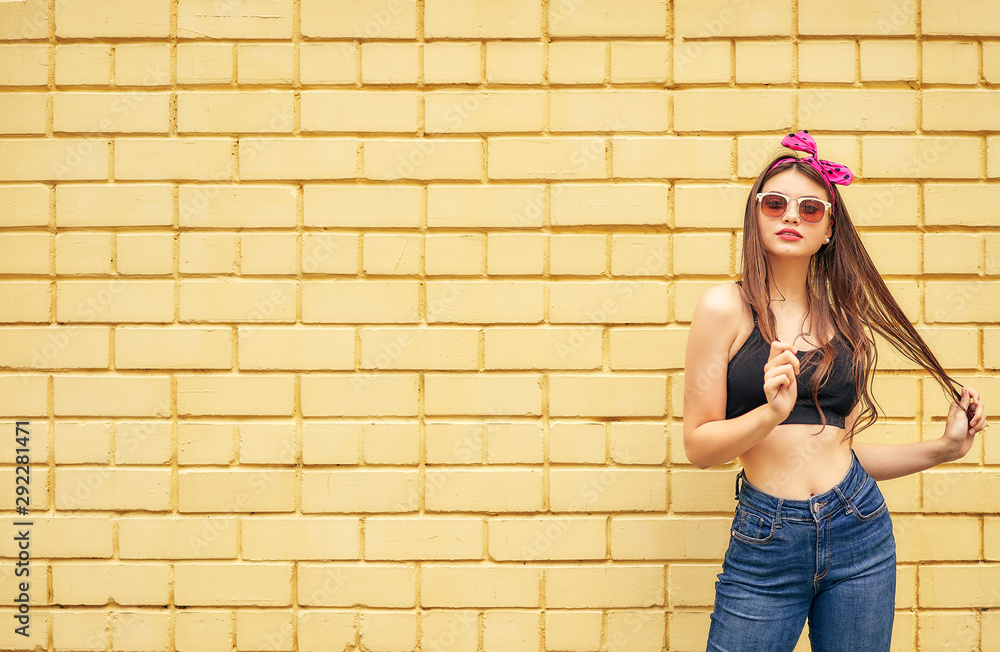 cheerful woman with a bow and a black tank top emotionally posing in sunglasses and jeans on a background of a yellow brick wall. copy space.