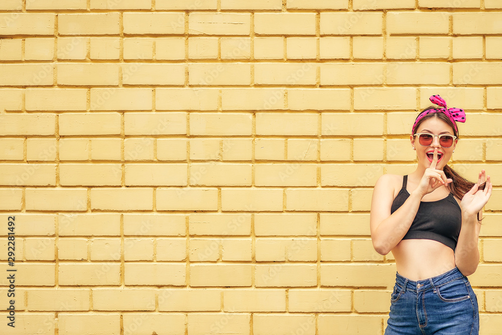 cheerful girl with a bow and a black tank top emotionally posing in sunglasses and jeans on a background of a yellow brick wall. copy space.