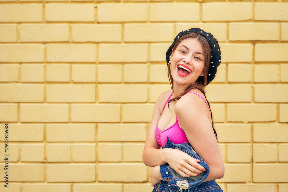 a cheerful girl in a beret and a denim jacket in her hands emotionally poses against a yellow brick wall in a red tank top and jeans. copy space.