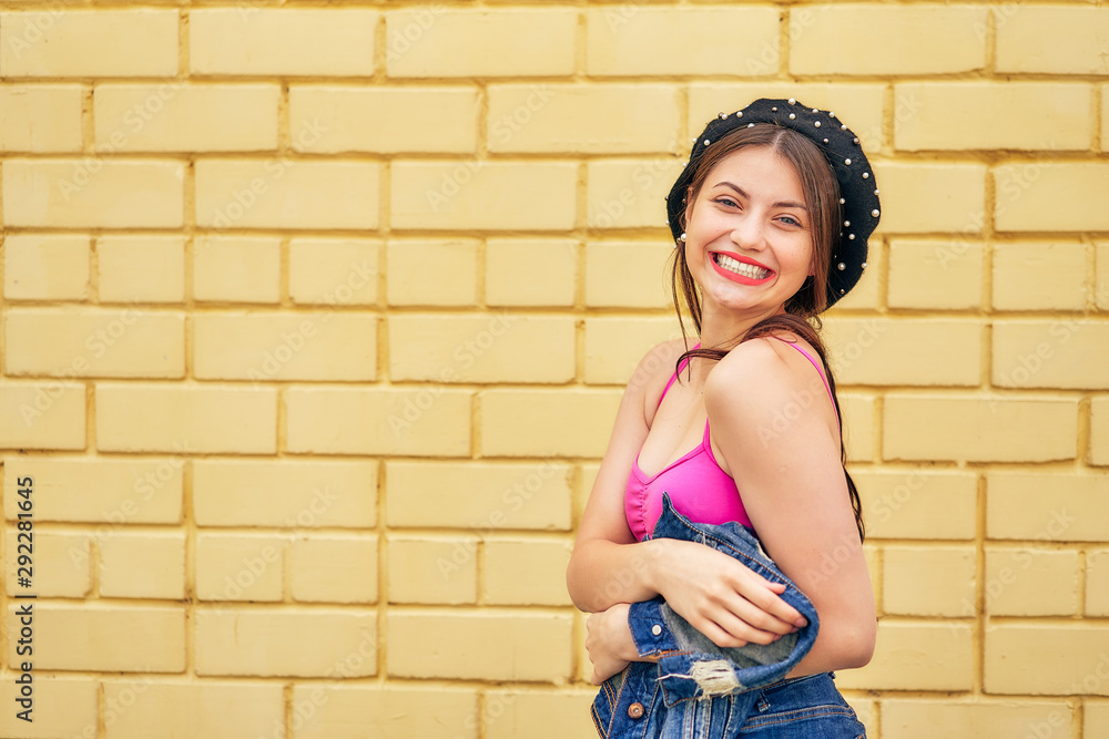a cheerful girl in a beret and a denim jacket in her hands emotionally poses against a yellow brick wall in a red tank top and jeans. copy space. a cheerful girl in a beret and a denim jacket in her hands emotionally poses against a yellow brick wall in a red tank top and jeans. copy space.