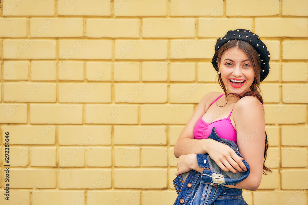 a cheerful girl in a beret and a denim jacket in her hands emotionally poses against a yellow brick wall in a red tank top and jeans. copy space. a cheerful girl in a beret and a denim jacket in her hands emotionally poses against a yellow brick wall in a red tank top and jeans. copy space.