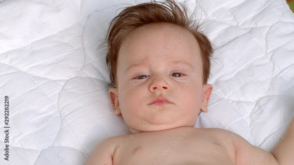 White little child with dark thick hair and dark eyes is lying topless on the blanket on open air and observes the area. It is moving head and fingers. It raises left and right hand White little child with dark thick hair and dark eyes is lying topless on the blanket on open air and observes the area. It is moving head and fingers. It raises left and right hand