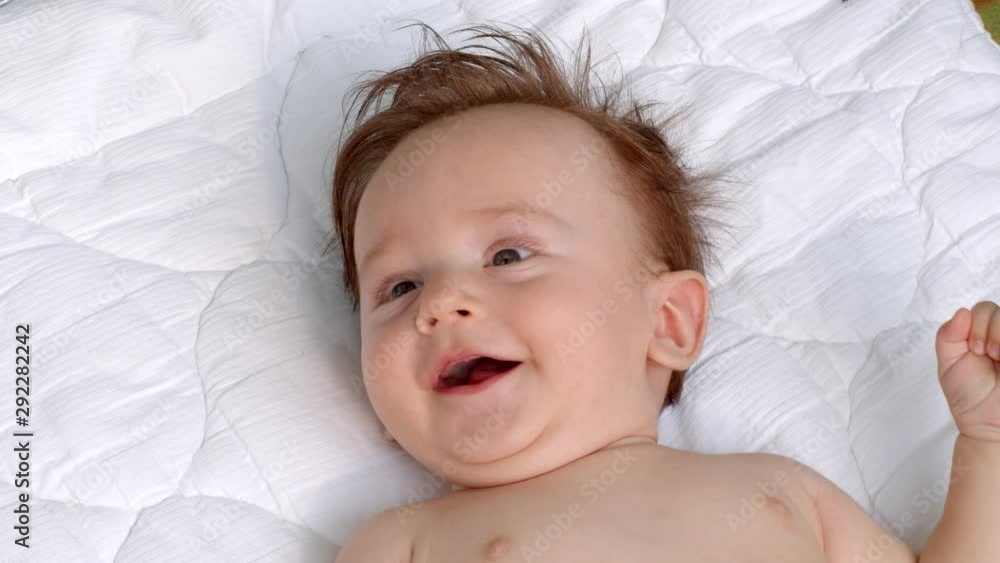 White smiling little child with dark thick hair and dark eyes is lying topless on the blanket on open air and observes the area. It is moving head and fingers. It raises left and right hand White smiling little child with dark thick hair and dark eyes is lying topless on the blanket on open air and observes the area. It is moving head and fingers. It raises left and right hand