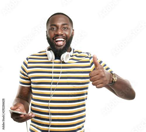 Happy African-American man with mobile phone and headphones showing thumb-up gesture on white background
