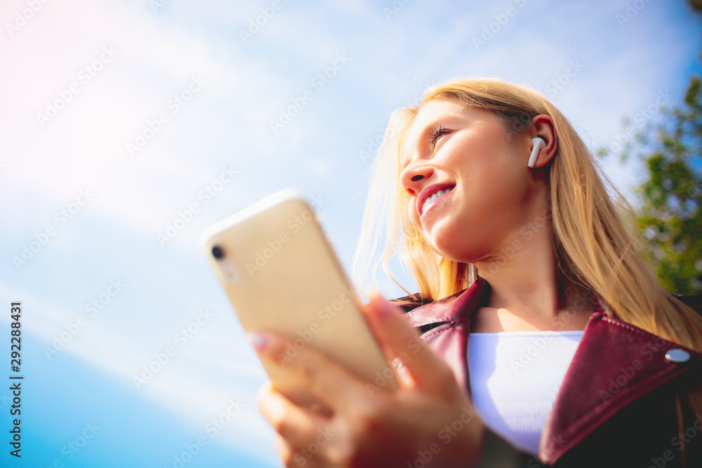 Young beautiful smiling adult Caucasian girl walking outdoor in the park having chat communication with wireless headphones mobile reading talking listening using with her hand on sunset at summer Young beautiful smiling adult Caucasian girl walking outdoor in the park having chat communication with wireless headphones mobile reading talking listening using with her hand on sunset at summer
